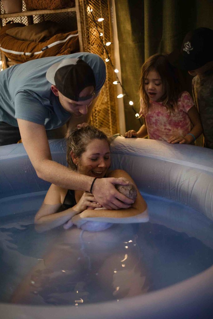 Woman in birth tub holding new baby next to husband and other children