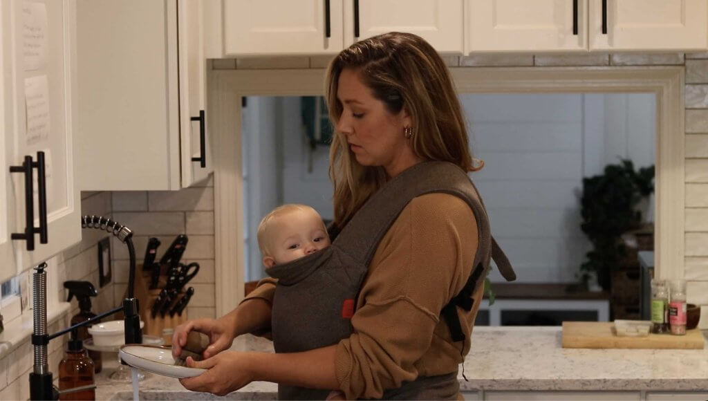 woman washing dishes while wearing her baby in a baby carrier
